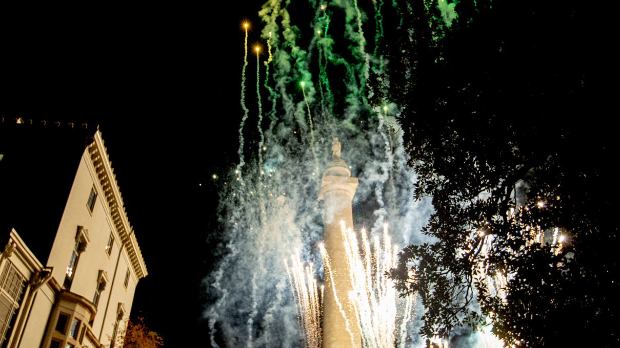 green and orange fireworks explode around the Baltimore Washington Monument with Walters Museum in the distance