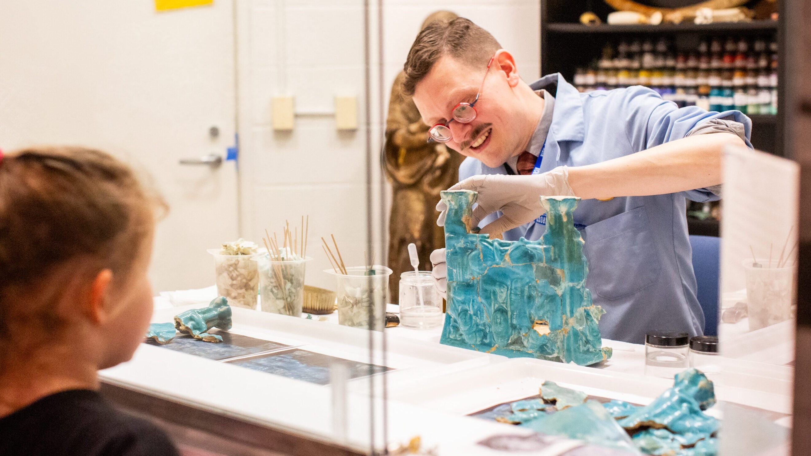 An art conservator works on an object while a visitor observes