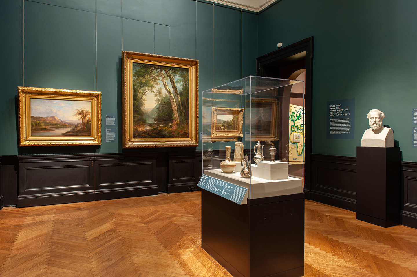 In-gallery view of Selections from the North American Collection: People and Places, which shows art objects in a case and paintings with a doorway behind.