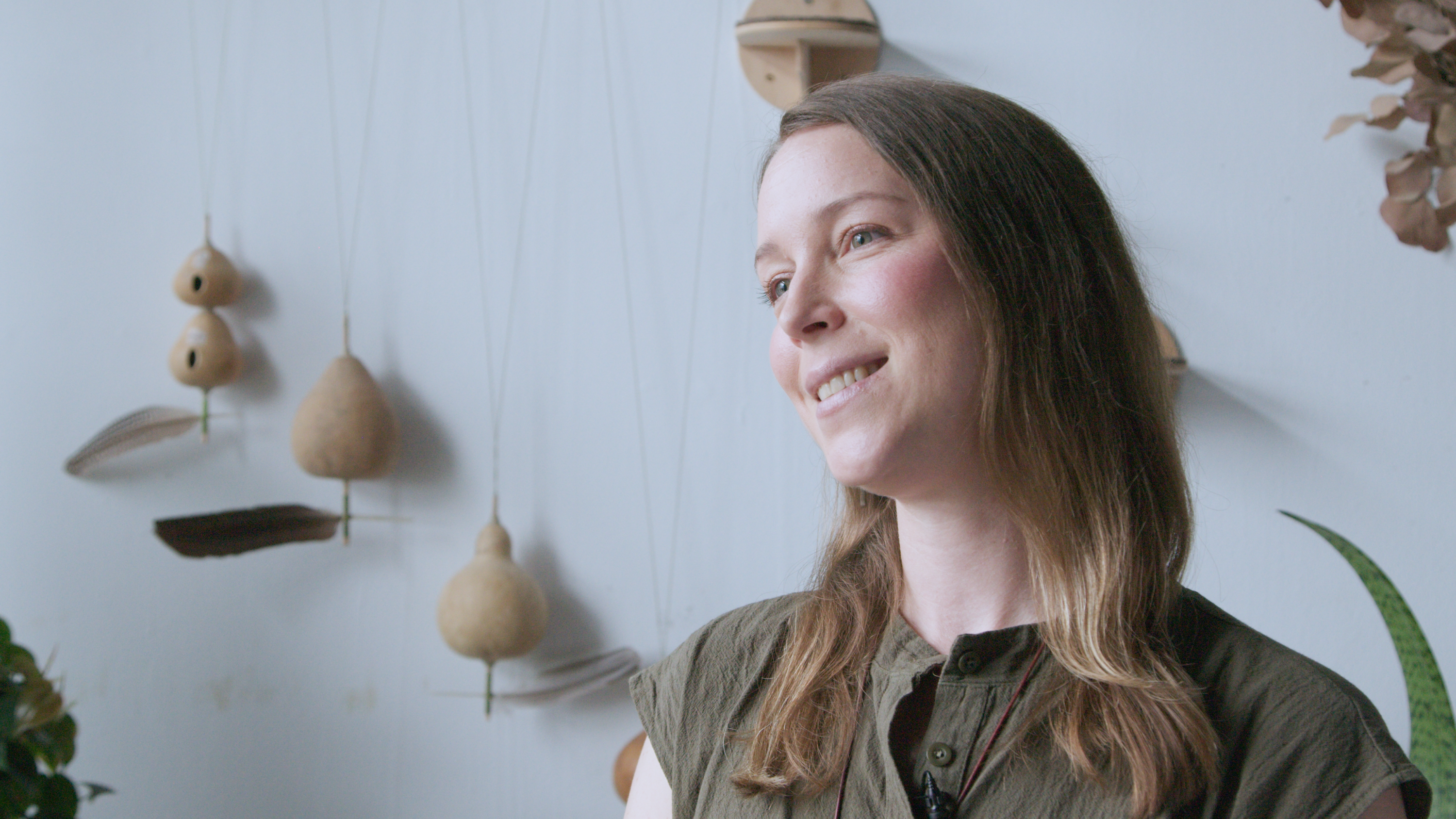 A woman in an olive green top smiles at the camera, while ocarinas hang on the wall behind her.