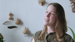 A woman in an olive green shirt smiles at the camera, while ocarinas hang on the wall behind her.