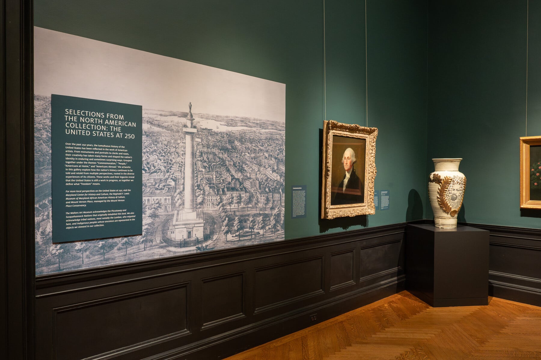 Gallery view of the United States at 250 showing a large wall graphic of the Washington Monument in Baltimore, an introductory text, a painting, and a vase
