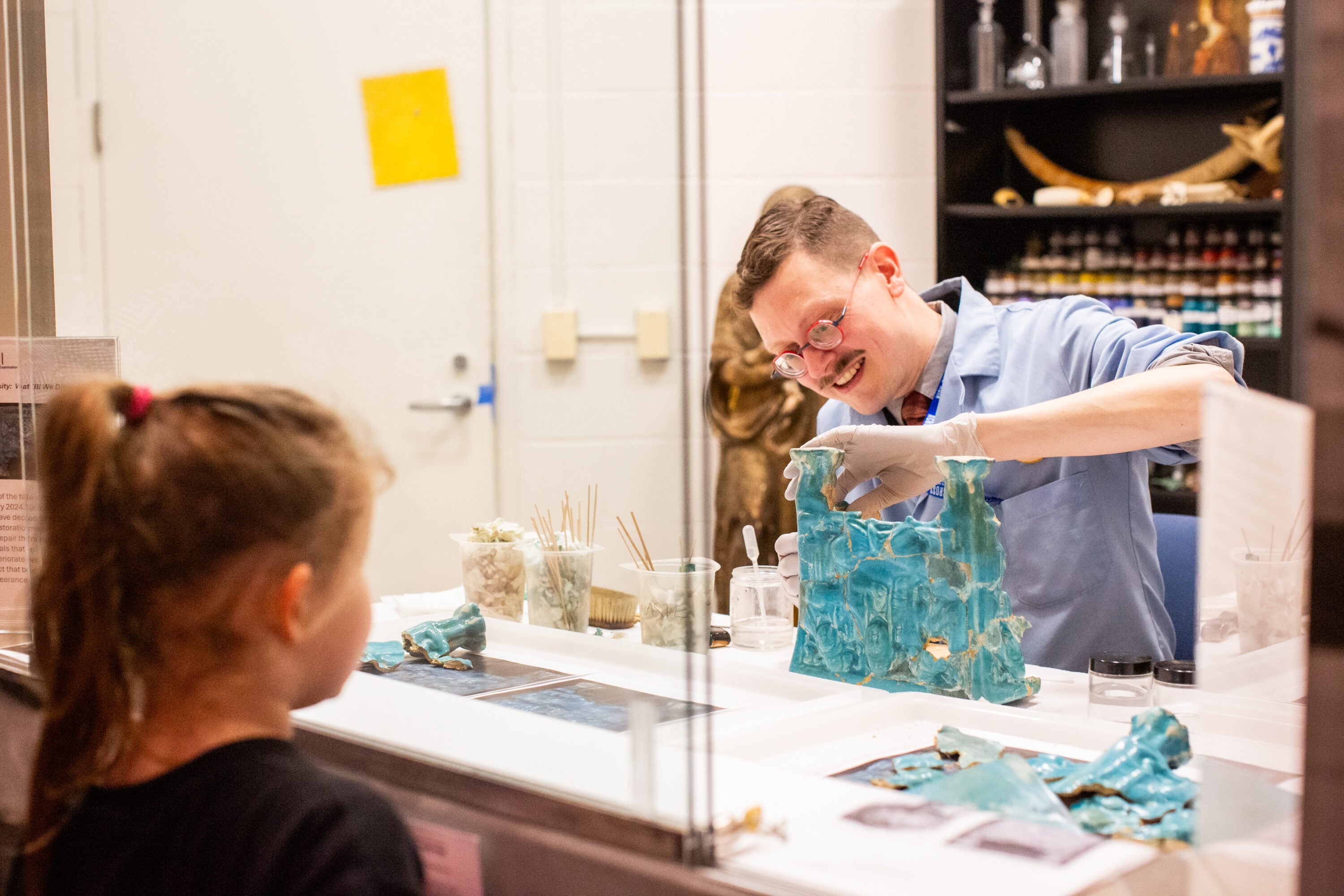 An art conservator works on an object while a visitor observes